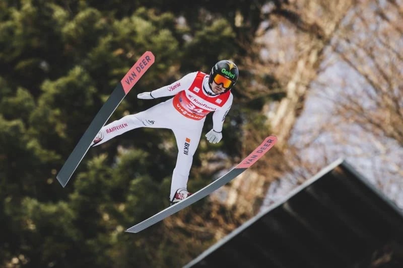 Skispringer Sandro Hauswirth in der Flugphase, die Ski in V-Stellung vor einem Waldhang